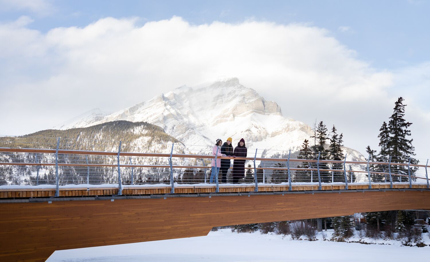 Three women stand on the Nancy Pauw bridge with Cascade Mountain behind them in Banff National Park.